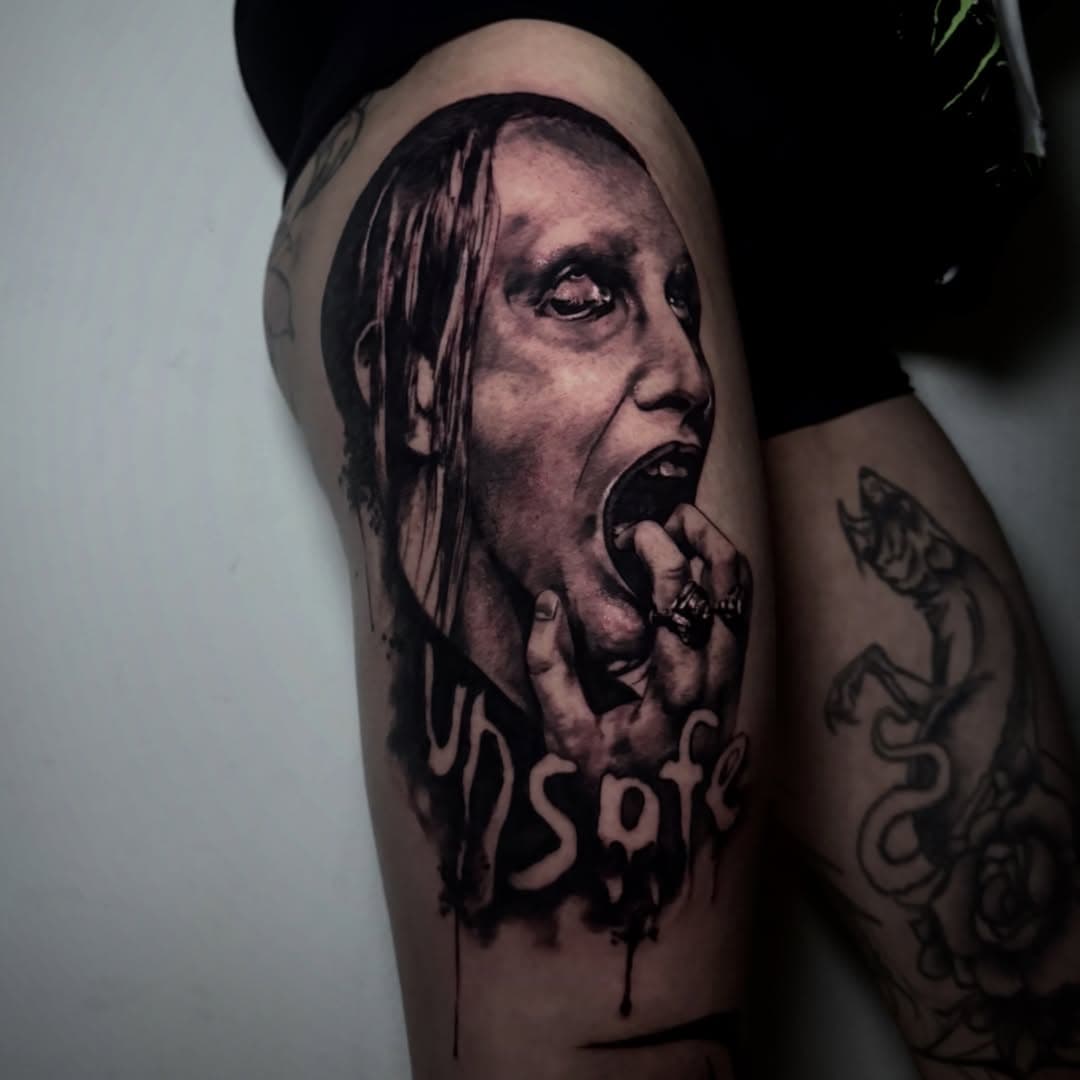 Black-and-grey realism portrait on the thigh with long hair, an open mouth and dripping lettering beneath it.