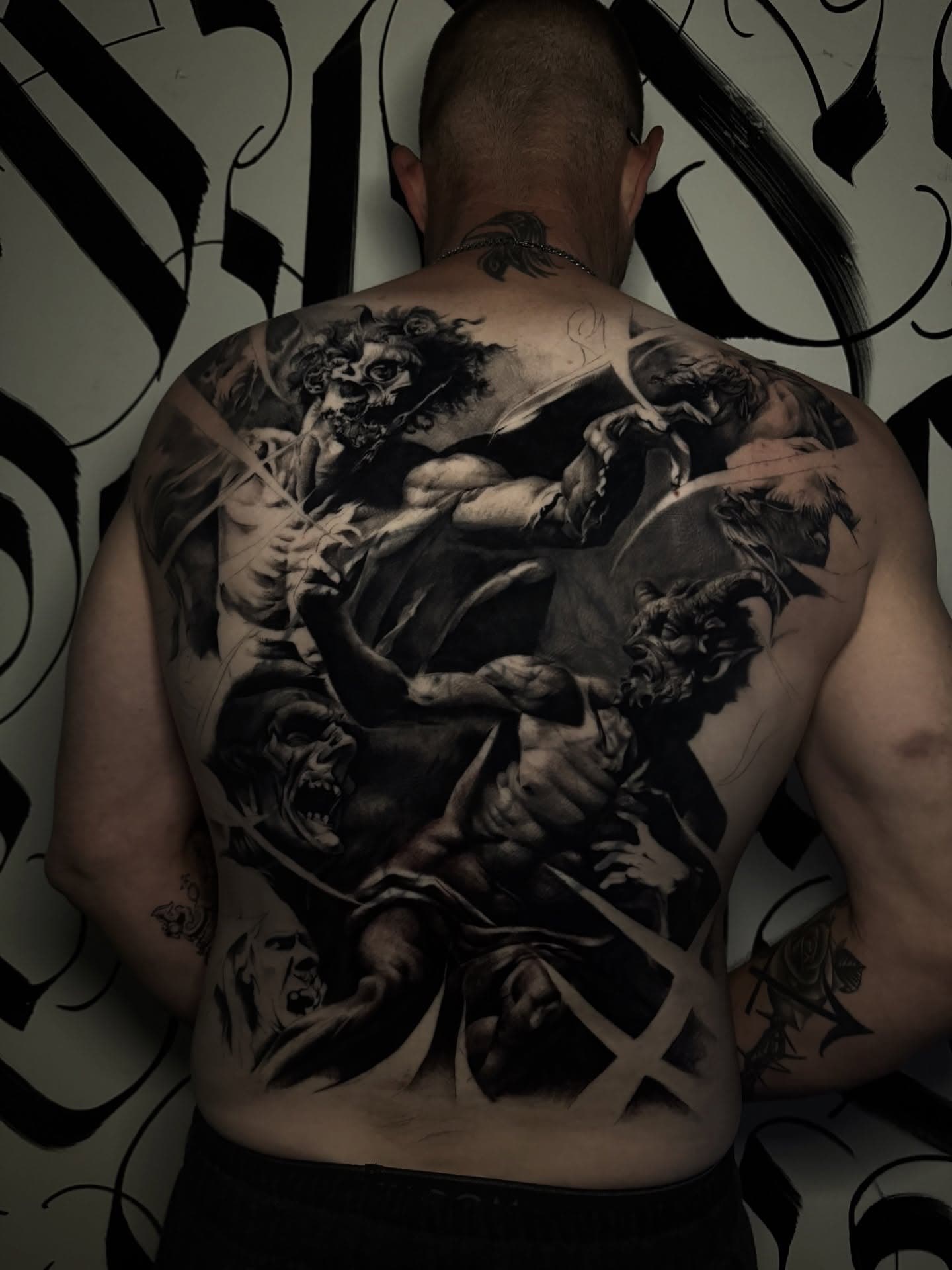 Full back tattoo of falling figures and snarling creatures in dense black and grey, built across the whole back and shoulders.