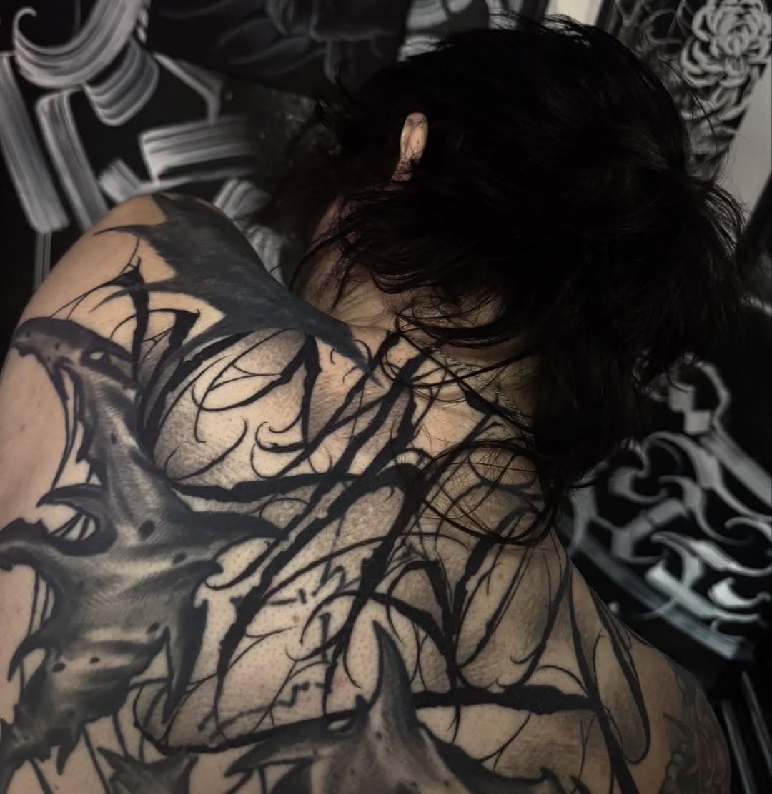 Dense gothic lettering and thorn-like blackwork covering the upper back and shoulder, shot in low light.